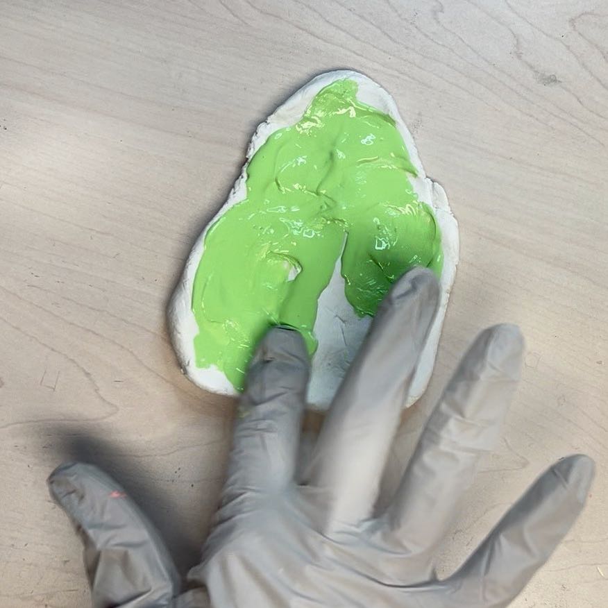 woman's gloved finger tips are spreading thee green paint over thee surface of the clay
