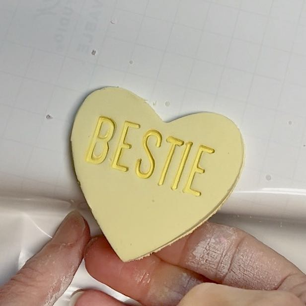 woman's fingers are holding a completed cllay heart with the word "BESTIE" stamped on it.