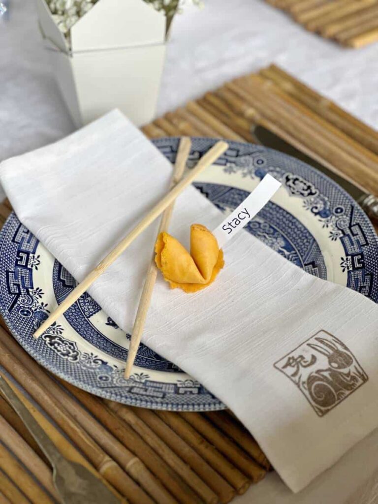 placesetting for a Chinese dinner party with a name coming out of a fortune cookie with chopsticksss and a napkin with a chinese symbol stamped on it.