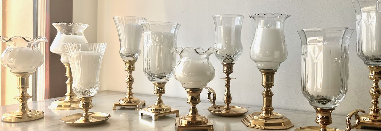 wide closeup of ten mismatched hurricane canddle holders in an assortment of brass candlesticks