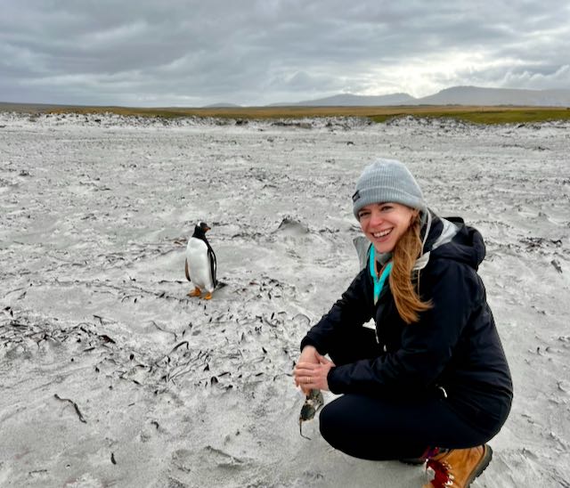 woman in winter garb kneeling on a grey sand beach close to a penguin