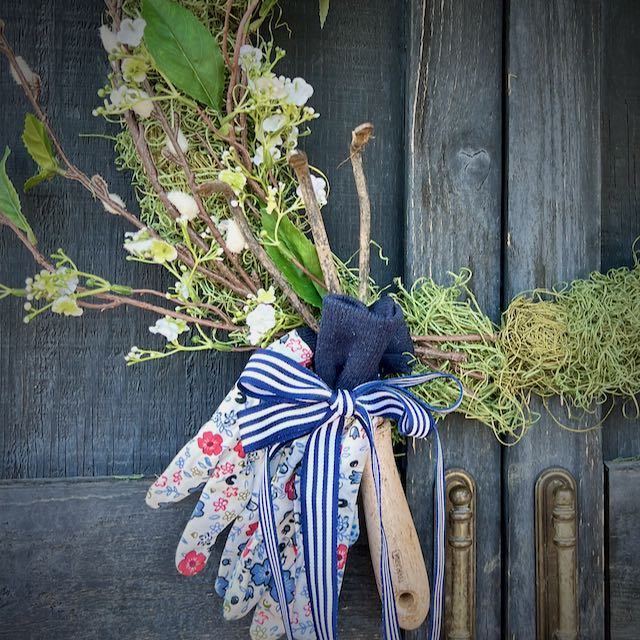 closeup of a pair of garden gloves, and old hand digger and some spring branches on a moss wreath on garden shed doors