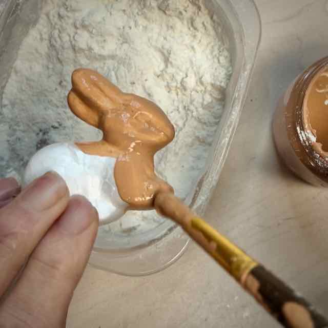 woman's fingertips are holding the back end of a foam bunny and a paint brush is painting the front end