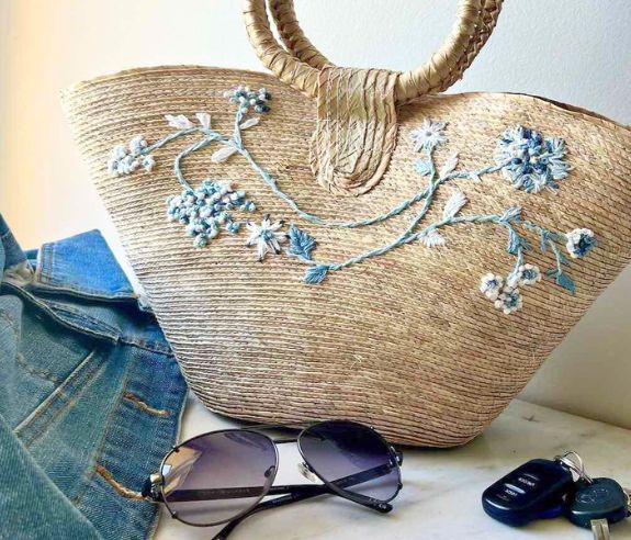 Embroidered straw handbag is sitting next to a denim jacket, sunglasses and keys