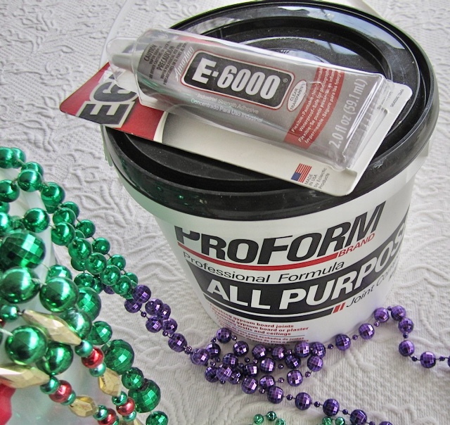 view of a pkg of E-6000 adhesive on top of a bucket of joint compound beside a pile of mardi gras beads
