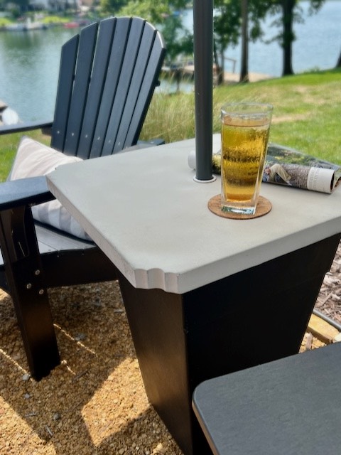 view of thee finished umbrella side table with concrete top holding a glass of iced tea and a magazine with lawn and lake in background