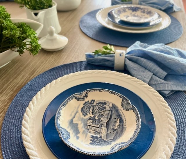 closeup of a blue and white table setting on round blur straw placemats with chambray napkins and white servings dishes with garden clippings for a centerpiece