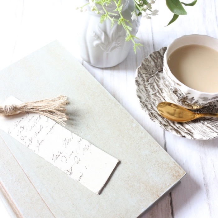 Tassel made of twine is attached to a bookmark and resting on top of a book beside a cup of coffee and sweet flowers