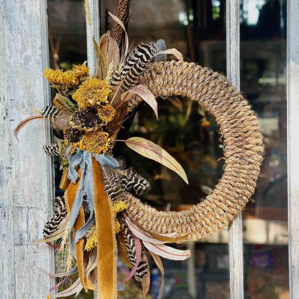 Fall wreath made of rope and embellished with dried flowers, leaves, feathers and ribbons is hanging on a glass door window