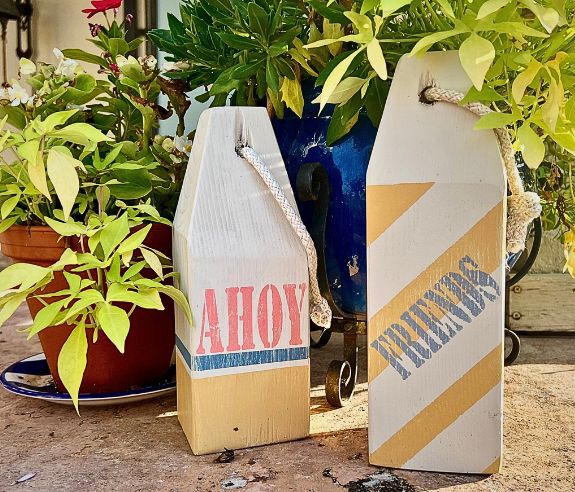 Two Wooden buoys that say Ahoy and Friends are on a stone ledge with potted plants behind