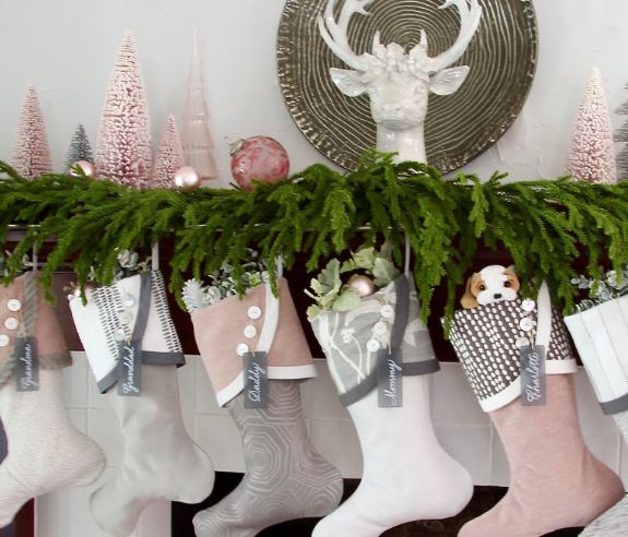 A Christmas mantel decorated with lush green garland and pastel-colored stockings. Each stocking is filled with small gifts and greenery, and labeled with names. Subtle pink Christmas trees and a decorative white deer head adorn the mantel above.