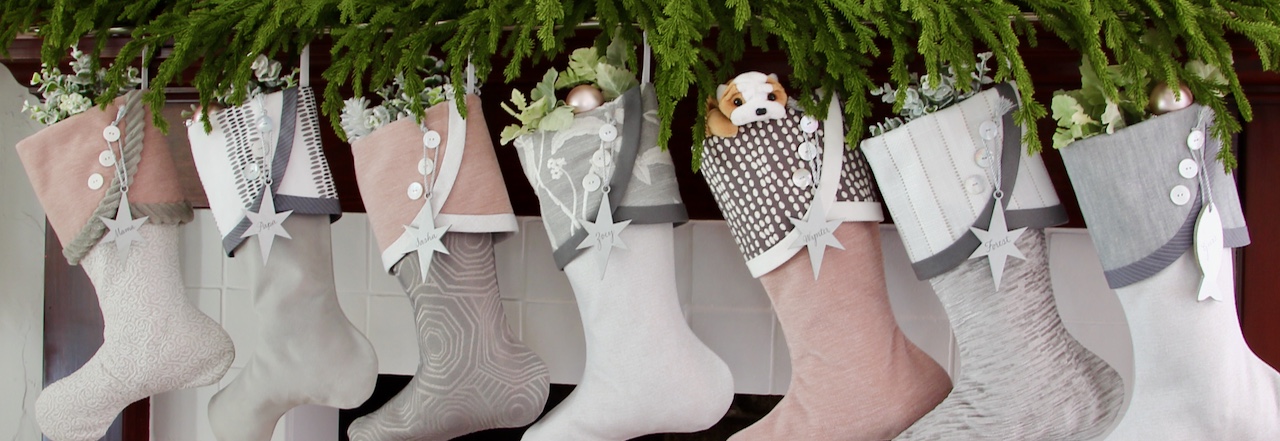 A row of Christmas stockings in various neutral tones hangs from a mantel adorned with greenery, reflecting the elegant charm of South House Designs Shop. Each stocking has decorative buttons or stars, and one stocking contains a small, brown and white plush dog. White flowers peek out of several stockings.