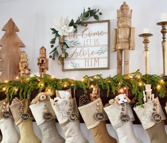 A cozy Christmas mantel display with stockings labeled with names hanging below. Above, there are nutcrackers, a wooden tree, candles, and a framed sign that reads "Oh Come Let Us Adore Him," adorned with greenery and lights.