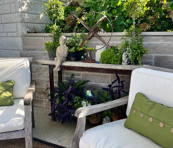 A cozy outdoor patio features white cushioned chairs with green accent pillows. A stone wall backdrop is adorned with lush greenery, a vintage sun dial, potted plants, and a small statue, creating a tranquil garden setting.