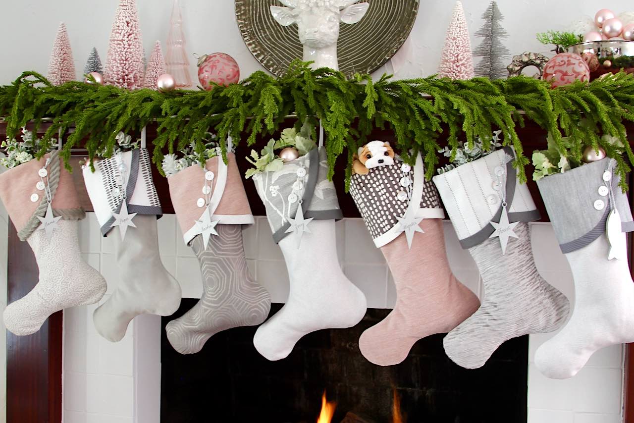 A fireplace adorned with a festive garland and seven 2023 Christmas stockings in pink, gray, and white tones. Each stocking boasts a unique design, with one holding a stuffed dog toy. The mantel is beautifully decorated with festive ornaments.