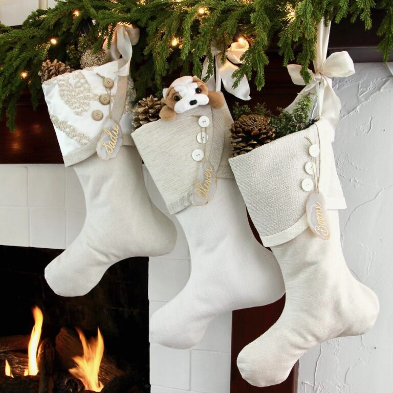 Three cream-colored Christmas stockings hang over a fireplace with a lit fire. Each stocking is adorned with buttons, pinecones, and personalized name tags. A stuffed dog peeks out of the middle stocking. Greenery decorates the mantel.