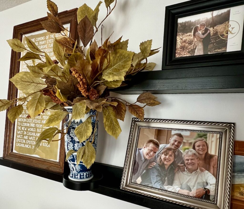 A wall with a DIY picture ledge displays dark shelves holding framed photos and a vase with autumn leaves. One photo shows a group of five people smiling, while another captures a couple embracing outdoors. A framed text piece is also elegantly positioned on the ledge.