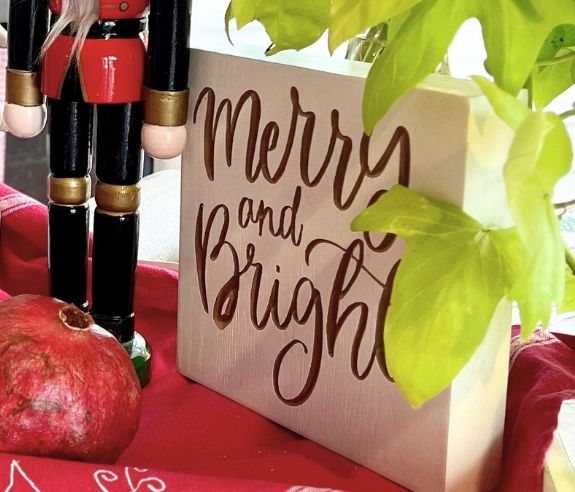 A decorative scene featuring a wooden block with the words "Merry and Bright" in cursive, a nutcracker figurine dressed in red and black, a pomegranate, and green leafy plants on a red cloth.