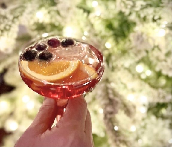 A hand holds a transparent cocktail glass filled with a pink drink, garnished with orange slices and berries. In the background, blurred, lit-up Christmas decorations can be seen.