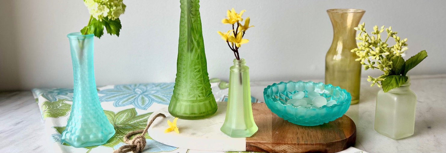 Various vases and a bowl serve as stunning bud vase centerpieces on the table, featuring green and blue glass. Each vase holds flowers or leaves, complemented by a wooden board and a floral-patterned cloth.