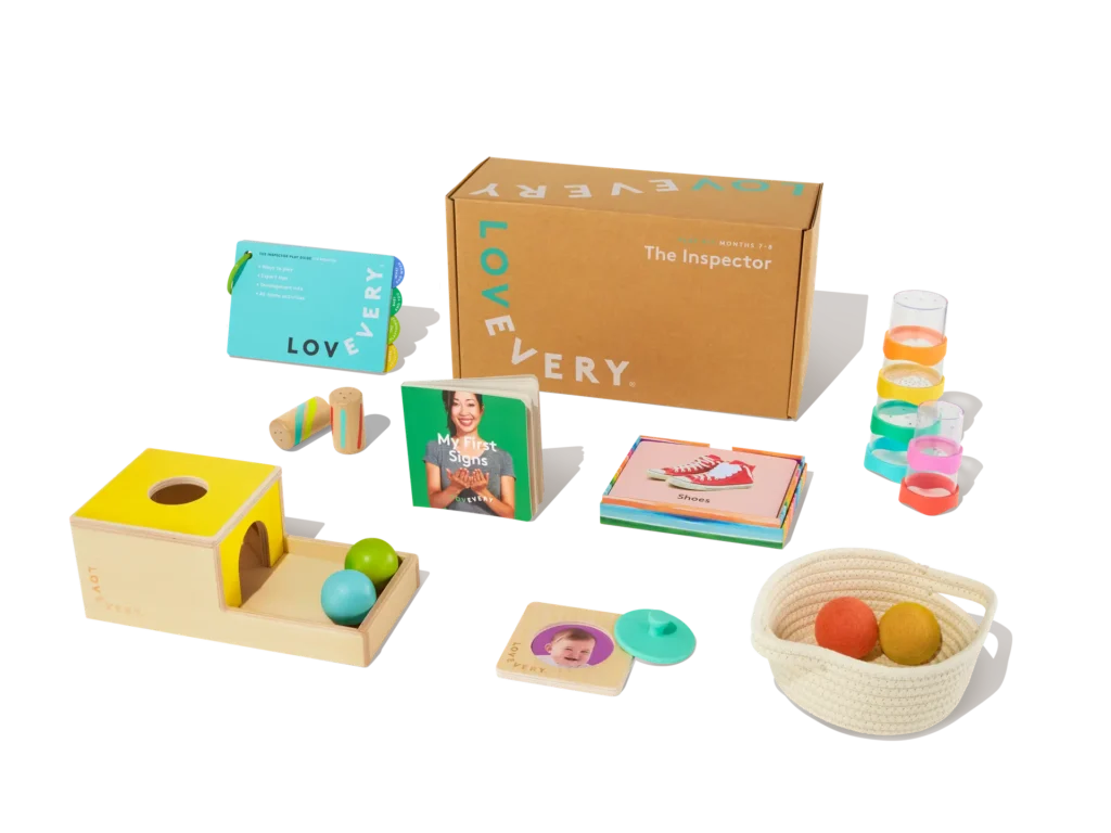 A collection of baby toys and books is displayed, including a wooden box with colorful balls, a board book, stacking cups, and a basket with balls. A cardboard box labeled "The Inspector" by Lovevery is in the background.