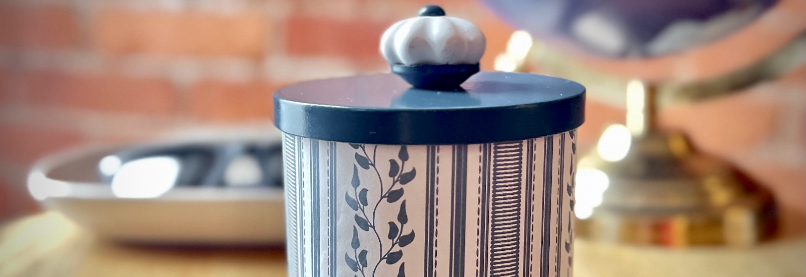 A decorative canister, repurposed from an old candle jar, features a striped and leaf pattern on a wooden table. It has a dark lid with a white knob. In the blurred background, there’s a brass lamp and a wall with a brick design.