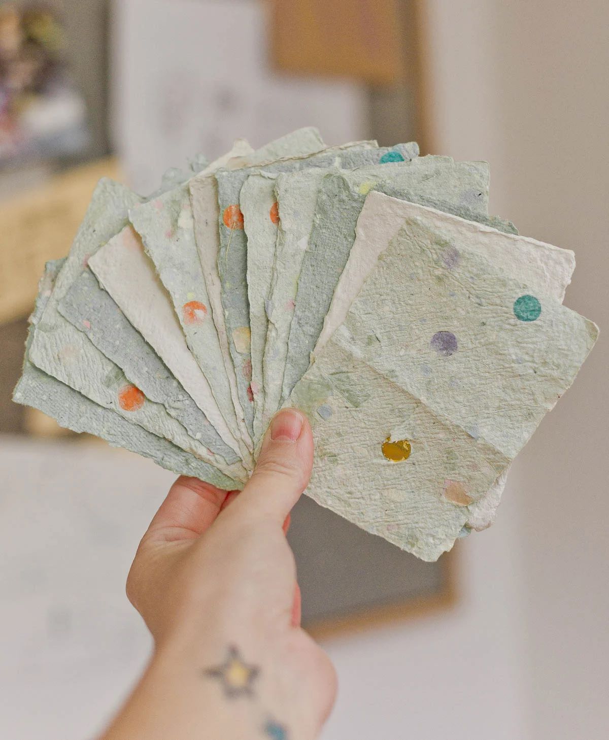A hand holds a fan of handmade paper sheets. The sheets are light green with textured surfaces and scattered colorful dots. A blurry background suggests an indoor setting with papers on a wall.