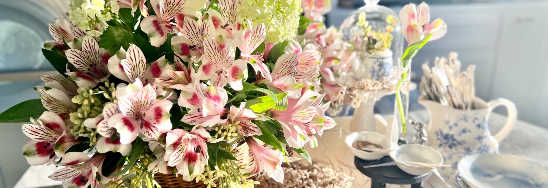 A vibrant floral arrangement, mixing fresh and artificial pink and white flowers with green leaves, is displayed in a wicker basket. Nearby, teacups, saucers, and utensils complement the decorative blue and white theme in the softly lit room.
