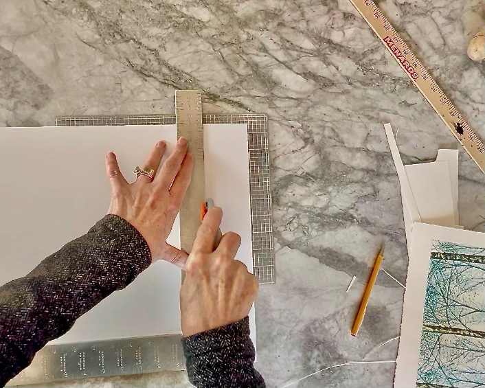A person uses a ruler and a cutter to trim a sheet of paper on a marble surface. A pencil, additional cut paper pieces, and measuring tools are scattered around. The person is wearing a dark speckled sweater.