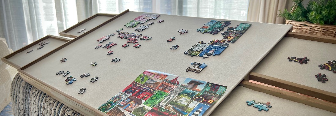 A partially completed puzzle with colorful pieces is spread across a large board with side trays near a patterned ottoman. The background shows sheer curtains and potted plants in soft lighting.