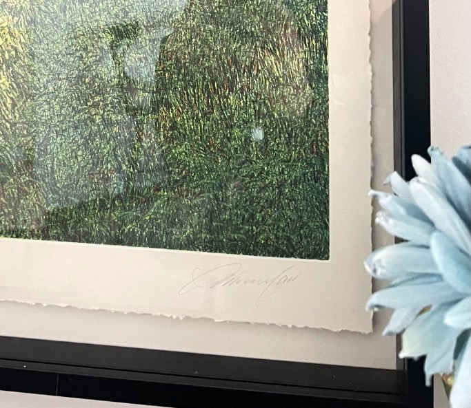 A close-up of a framed artwork with green textured patterns, resembling foliage or vegetation. The frame's edge is visible, and a soft-focus blue flower is in the foreground on the right side of the image.