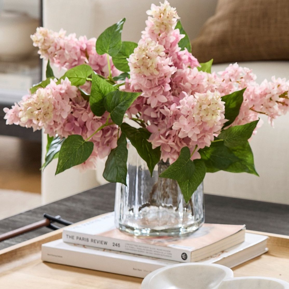 A vase with light pink hydrangeas, crafted using the best fake flower tips, sits on a stack of books on a wooden table. A brown pillow graces a white couch in the background, creating a cozy and elegant living room setting.