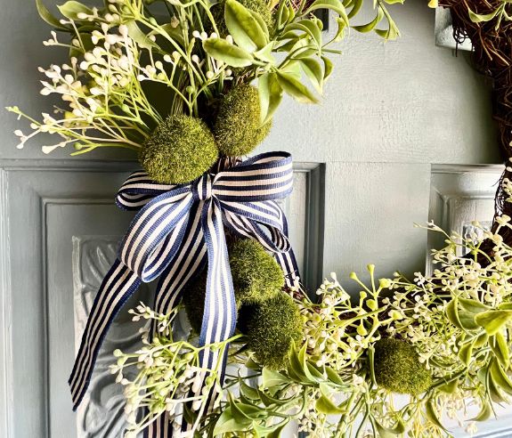 A close-up of a green wreath with clusters of small white flowers, mossy green balls, and a blue-and-white striped bow, hanging on a light blue door.