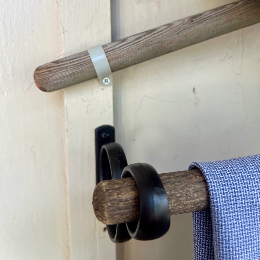A close-up of a wooden rod mounted on a wall with black metal brackets, holding a blue and white checkered towel and three black rings—perfect above a shelf to display or decorate candles.