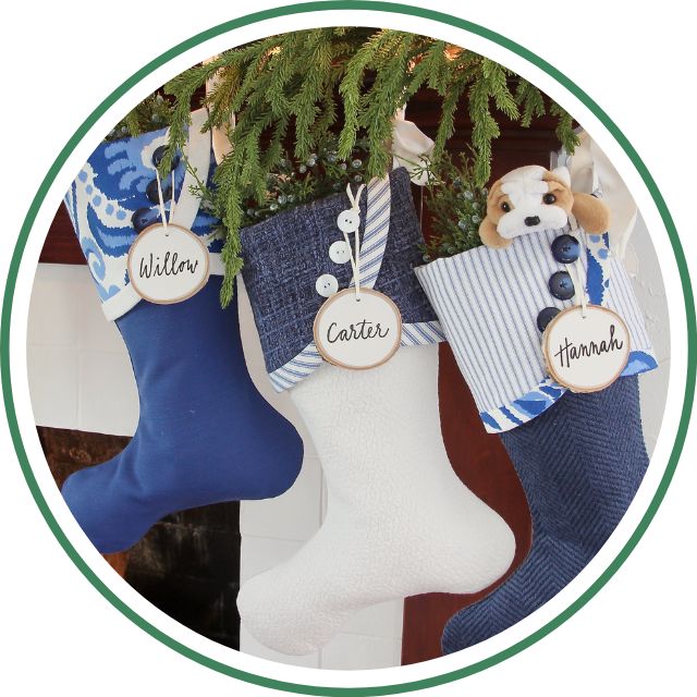 Three Christmas stockings in blue and white patterns hang from a mantel, each labeled with a wooden tag reading "Willow," "Carter," and "Hannah." A plush puppy peeks out of Hannah's stocking, offering a festive business opportunity for holiday décor.