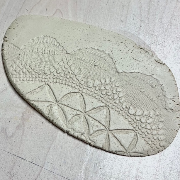 A flat, oval-shaped diy tissue holder made from clay features intricate patterns resembling lace and geometric shapes pressed into its surface, resting on a light wooden background.
