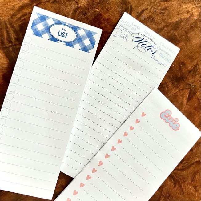 Three decorative notepads on a wooden surface: one with a blue checked header reading "the LIST," one titled "Notes" with words like "thoughts" and "ideas," and one with "errie" and pink hearts.
