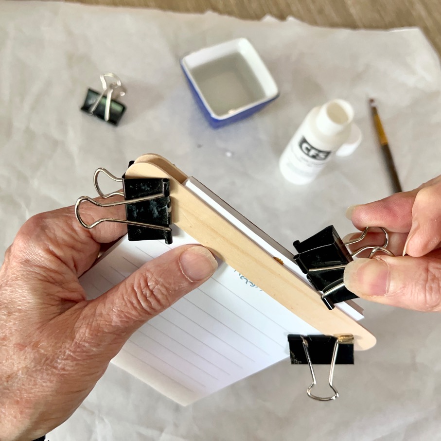 A person uses binder clips to hold together a stack of index cards with a wooden strip while preparing to glue the edge. Glue, a brush, and a small dish are in the background on a piece of parchment paper.