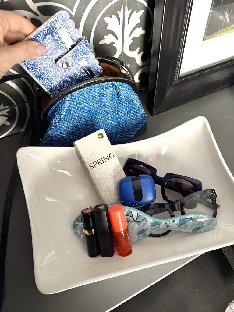 A hand places a blue-patterned wallet into a blue woven pouch. In front, a white tray holds sunglasses, perfume, lipsticks, earbuds in a blue case, and a small patterned hairbrush.