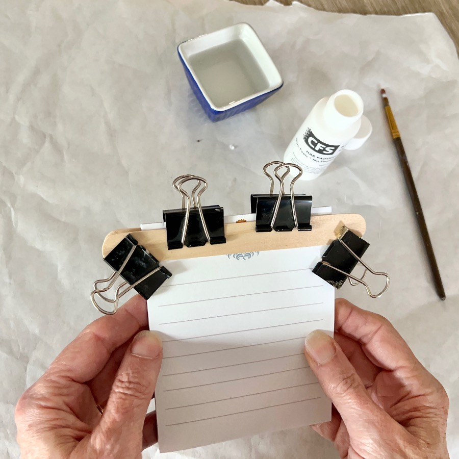 Two hands hold a stack of lined paper clamped by binder clips to a wooden stick. Nearby are a small dish of water, a bottle of adhesive, and a paintbrush on a white surface.