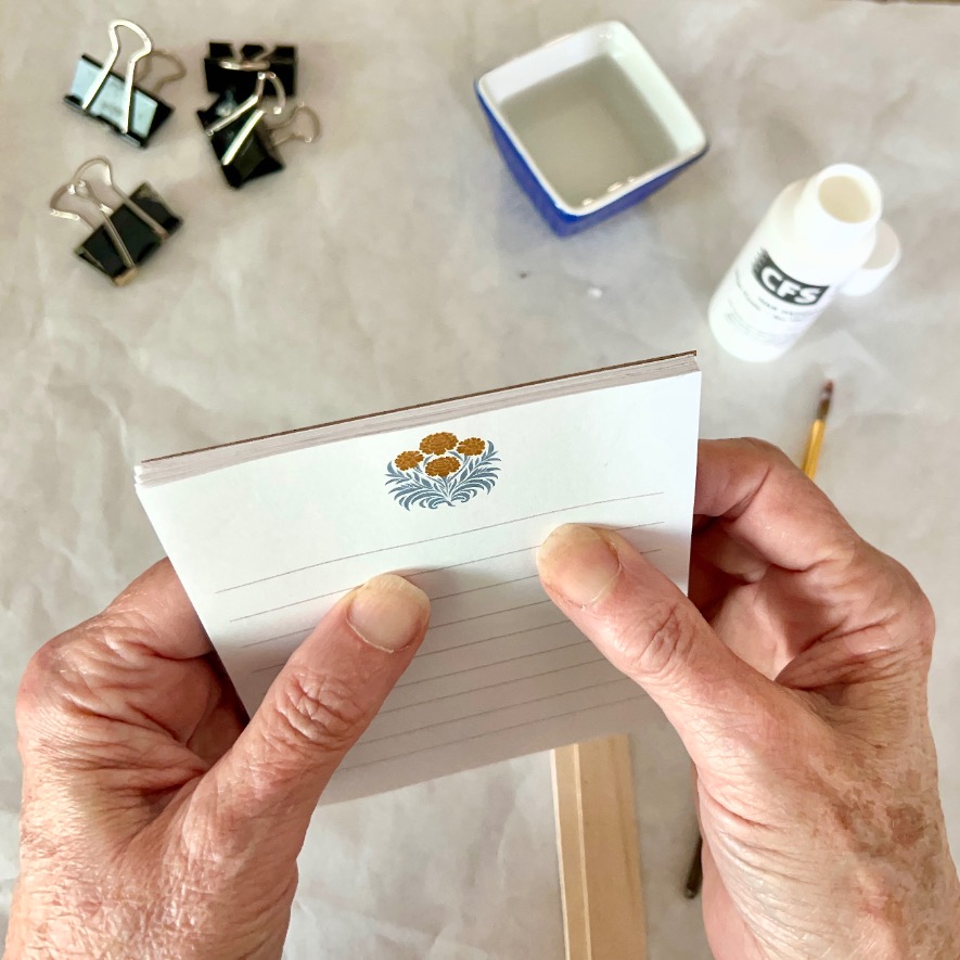 Two hands hold a small notepad with a floral design at the top. On the table are binder clips, a bowl of water, a bottle of glue, a paintbrush, and a wooden stick, suggesting a DIY notepad project.