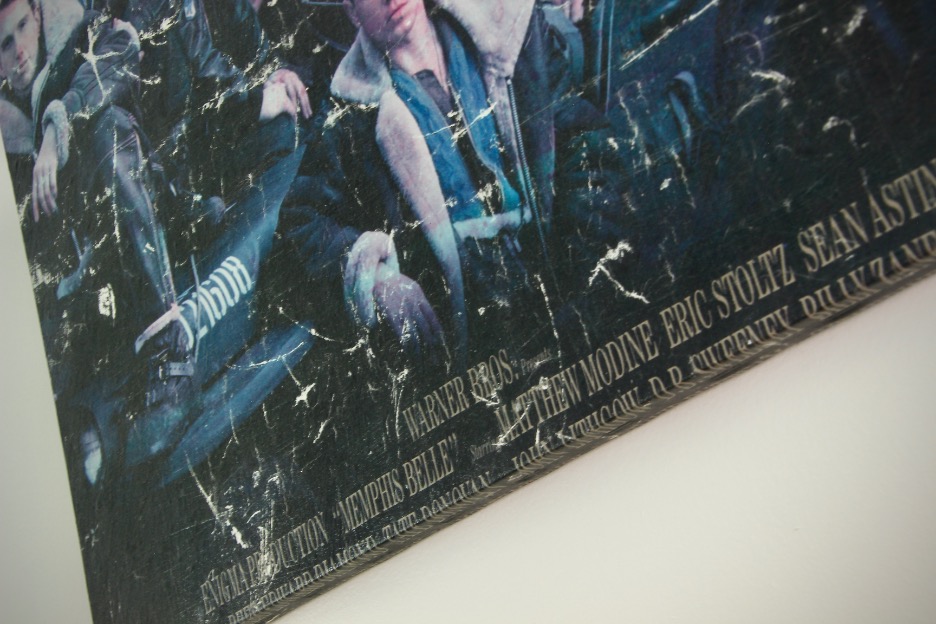 A close-up of vintage poster artwork shows a worn and scratched movie poster featuring a group of people, with visible credits at the bottom listing stars like Eric Stoltz and Sean Astin. The poster is mounted on a white wall.
