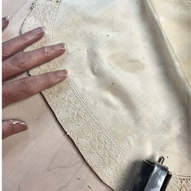 A hand with paint on the fingers touches the edge of a cream-colored fabric with lace trim, crafting a diy tissue holder. Part of a metal tool is visible in the lower right corner. The fabric rests on a light wood surface.