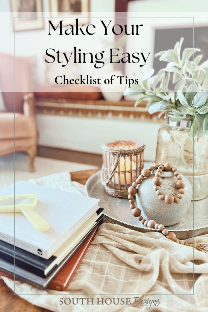 A cozy living room scene featuring a tray with a candle, wooden beads, and a plant on a beige ottoman—perfect inspiration to style your round coffee table. Stacked books and pastel note cards complete the look. “Make Your Styling Easy Checklist of Tips.”.