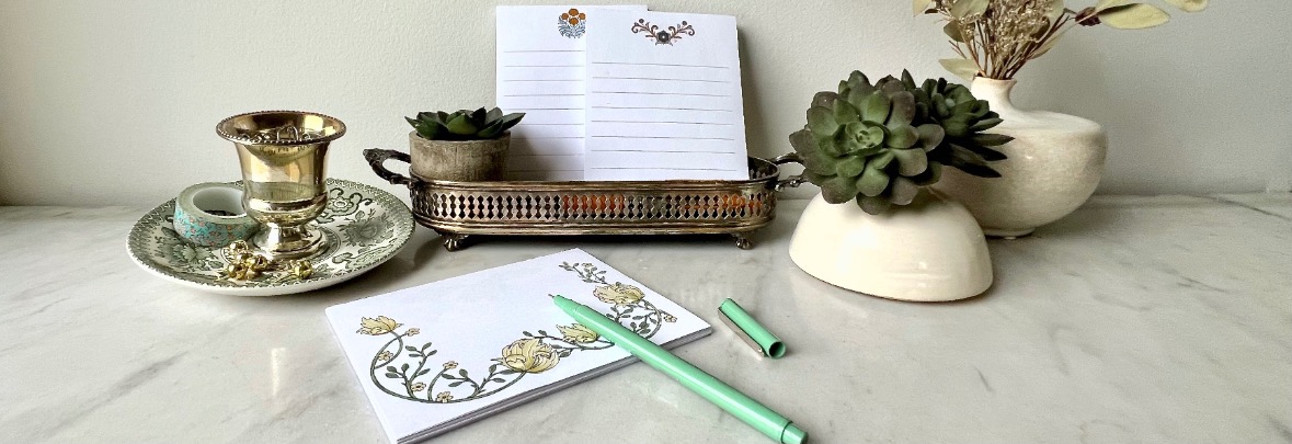 A marble desktop with succulents, a decorative cup on a saucer, a silver tray holding lined papers, and floral stationery with a green pen and pen cap placed nearby. A vase with dried flowers is in the background.