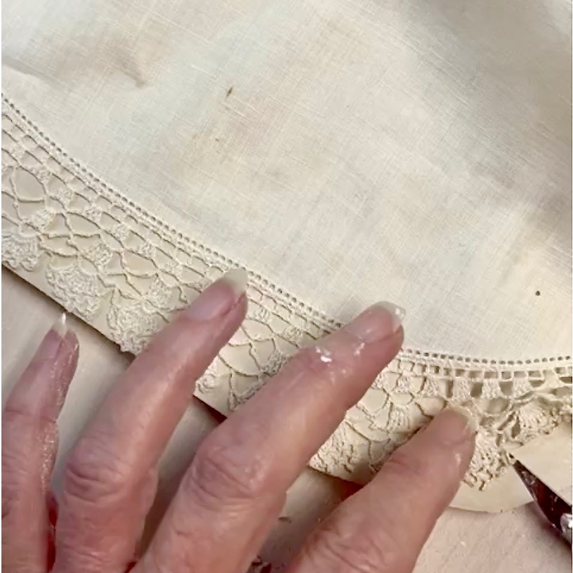A close-up of a hand with glue on the fingertips pressing lace trim onto the edge of a light-colored fabric diy tissue holder, with scissors partially visible in the corner.