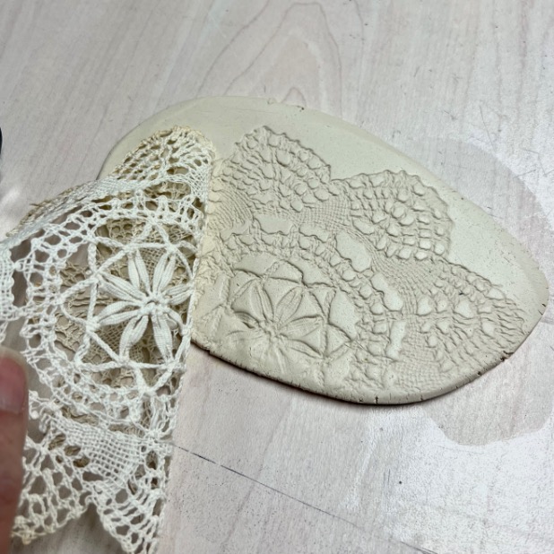 A hand lifts a piece of lace off a flat piece of clay, revealing an intricate lace pattern imprinted on the clay—perfect for crafting a unique diy tissue holder. The clay rests on a light wooden table.