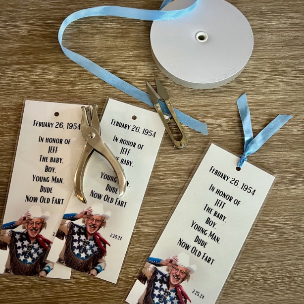 A table displays custom bookmarks with a photo of an older man in patriotic attire, text honoring him, a hole punch, blue ribbon, small scissors, and ribbon—creative inspiration for adult birthday party ideas.