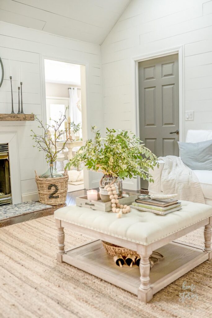 Bright, airy living room with a style round coffee table holding greenery, books, beads, and a candle. Neutral tones, a woven rug, wicker basket with branches, and light walls create a cozy, inviting space.
