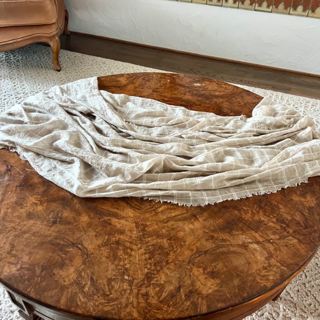 A beige, grid-patterned fabric is draped loosely over a style round coffee table in a living room with a textured white rug and a vintage chair in the corner.